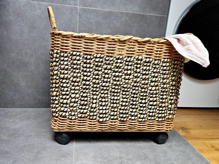 Pleciony kosz na pranie na kółkach wykonany z trawy morskiej w kolorze brązowym i czarnym. Produkt posiada solidne uszy i kółka ułatwiające przemieszczanie, pokazany na tle szarych płytek obok pralki z przewieszoną jasną tkaniną.