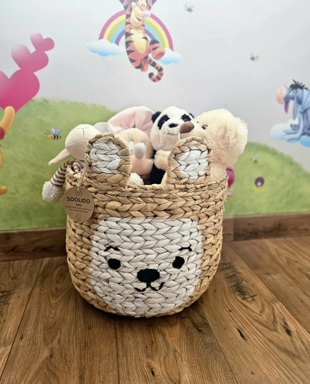 Pleciony kosz z hiacyntu wodnego w kształcie misia z uszami, wypełniony pluszakami, stojący na drewnianej podłodze w pokoju dziecięcym na tle kolorowej fototapety.