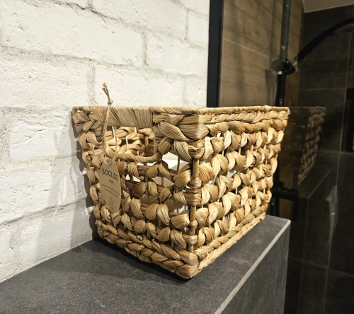 ostokątny kosz pleciony z grubych splotów hiacyntu wodnego w kolorze naturalnego brązu, ustawiony na ciemnoszarym blacie na tle białej ceglanej ściany. Kosz posiada wycięte uchwyty i ozdobną papierową etykietę marki Soolido.