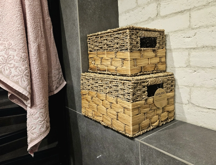 Dwa ozdobne kosze plecione z hiacyntu wodnego i trawy morskiej w stylu boho. Zestaw zamykanych pojemników z uchwytami ułożony na szarej półce w łazience obok białej cegły.