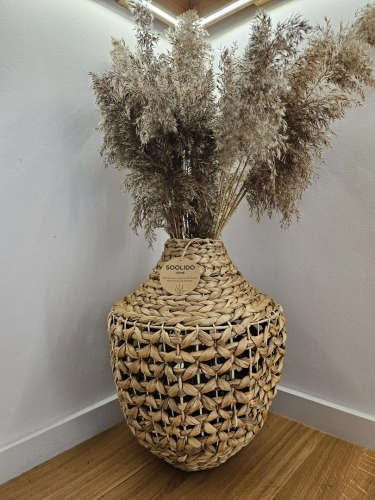 Duży pleciony wazon podłogowy z hiacyntu wodnego w stylu boho, wypełniony suszonymi trawami. Naturalny brązowy splot o dekoracyjnym ażurowym wzorze, stojący w rogu pokoju na drewnianej podłodze.