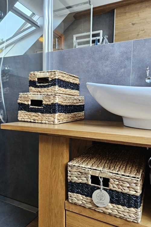 Trzy zamykane kosze z naturalnego hiacyntu wodnego w różnych rozmiarach. Design w poziome, brązowo-czarne pasy zintegrowany z otworami na dłonie. Kosze wyeksponowane na dębowej półce łazienkowej.