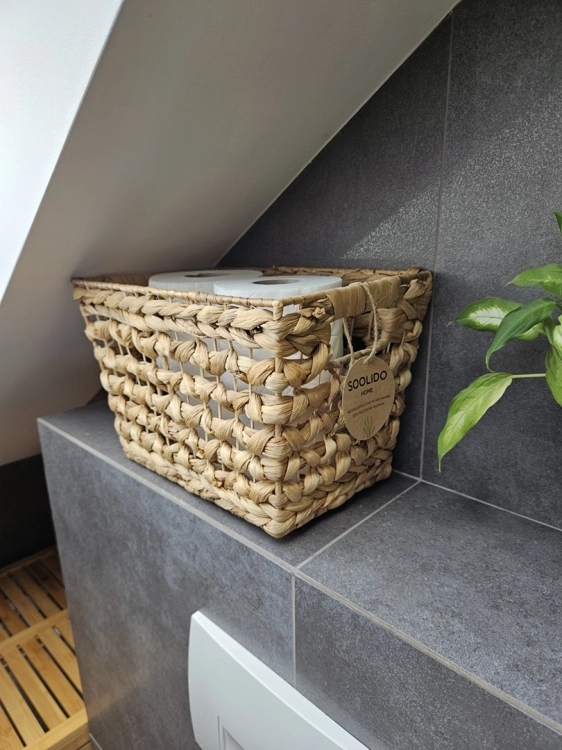 Pleciony kosz z hiacyntu wodnego w stylu boho, wypełniony papierem toaletowym. Ma prostokątny kształt, gruby splot i ozdobną zawieszkę Soolido. Stoi na szarych kafelkach w łazience.