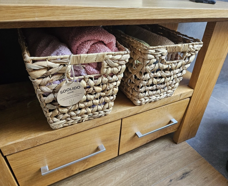 Brązowy kosz pleciony z hiacyntu wodnego w stylu boho, wypełniony ręcznikami, stojący na drewnianej szafce łazienkowej. Widoczna etykieta marki Soolido.