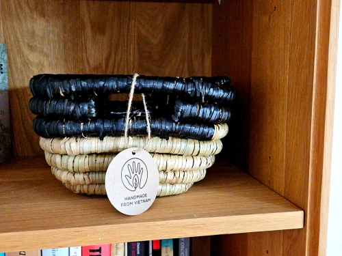 Pleciony kosz z trawy morskiej i hiacyntu wodnego w dwóch kolorach: naturalnym beżu i czerni. Produkt w stylu loftowym z okrągłą metką „Handmade from Vietnam”, wyeksponowany na drewnianym regale.