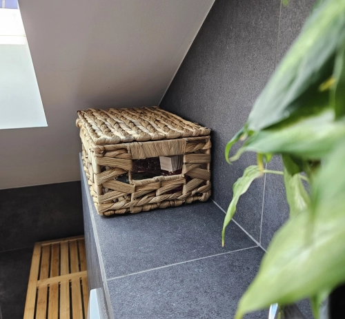 Prostokątny koszyk z hiacyntu wodnego z pokrywką, ustawiony na ciemnoszarej półce w łazience. Styl boho, naturalny splot, obok widoczna zielona roślina.