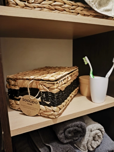 Pleciony koszyk z hiacyntu wodnego z pokrywką, beżowo-czarny, stojący na regale w łazience obok szczoteczek do zębów i ręczników. Styl boho, marka Soolido.