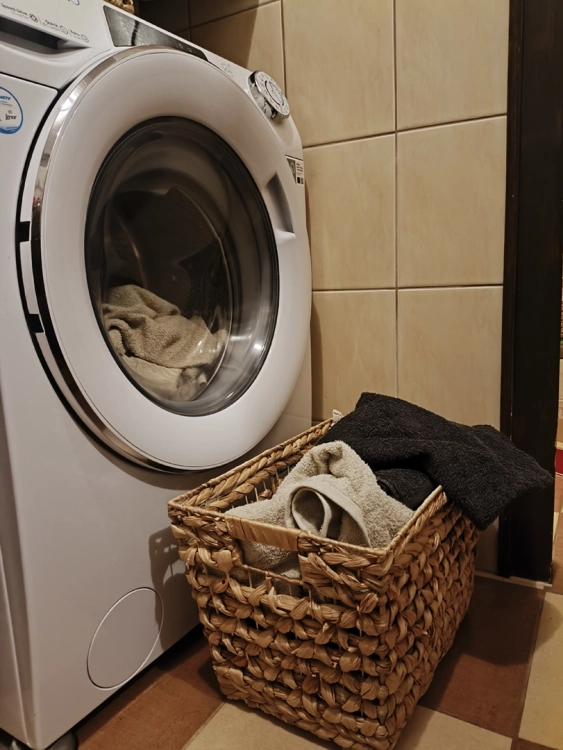 Prostokątny kosz z plecionego hiacyntu wodnego w stylu boho, wypełniony ręcznikami, stojący na podłodze w łazience obok białej pralki.