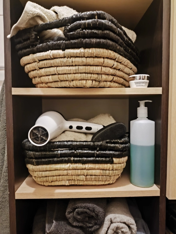 Dwa plecione kosze z trawy morskiej i hiacyntu w kolorze beżowo-czarnym, umieszczone na regale w łazience. Wewnątrz znajdują się ręczniki oraz suszarka do włosów. Styl boho.