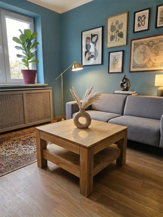 Wizualizacja salonu z kwadratowym stolikiem dębowym Witten w jasnym odcieniu dębu. Na stoliku wazon z trawą pampasową. W tle sofa, lampa i galeria zdjęć, oddające przytulną atmosferę.