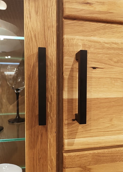 Zbliżenie na minimalistyczne, czarne metalowe uchwyty na drzwiach witryny dębowej Como 2, z widocznym naturalnym usłojeniem dębu i elementem przeszklonym.