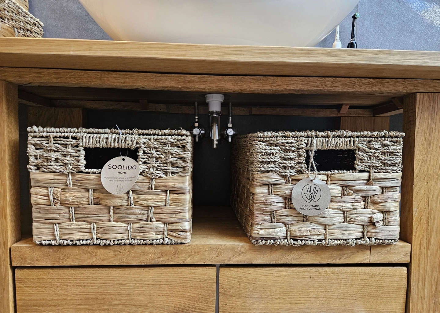 Prostokątne kosze plecione boho z hiacyntu i trawy morskiej. Widoczny dwukolorowy wzór i splot. Organizery łazienkowe pod umywalką na drewnianym regale. Soolido Home.